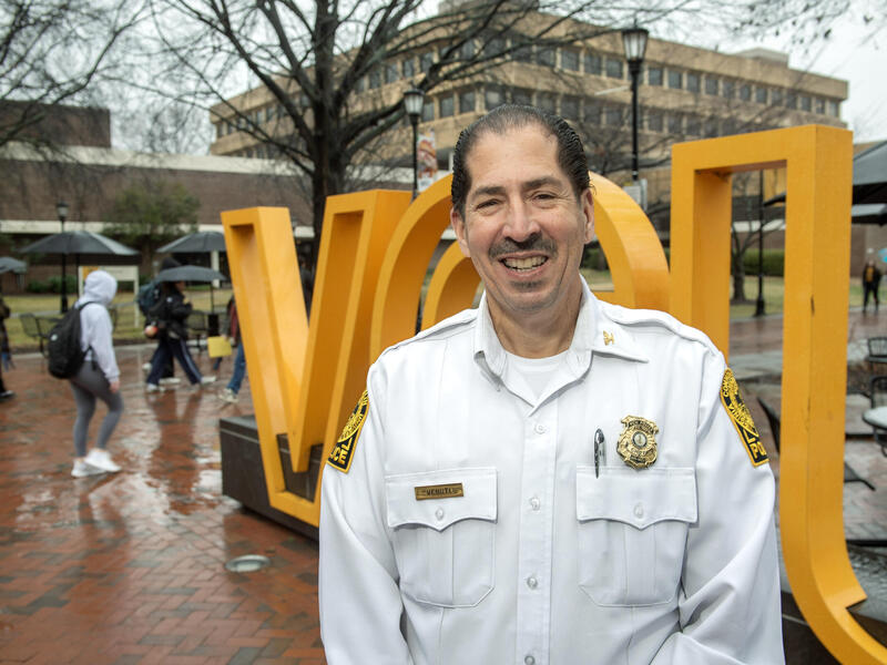 A photo of a man from the chest up. He is wearing a white police uniform shirt and a gold police badge. Behind him is a yellow sign that says \"VCU\" in large yellow letters. 