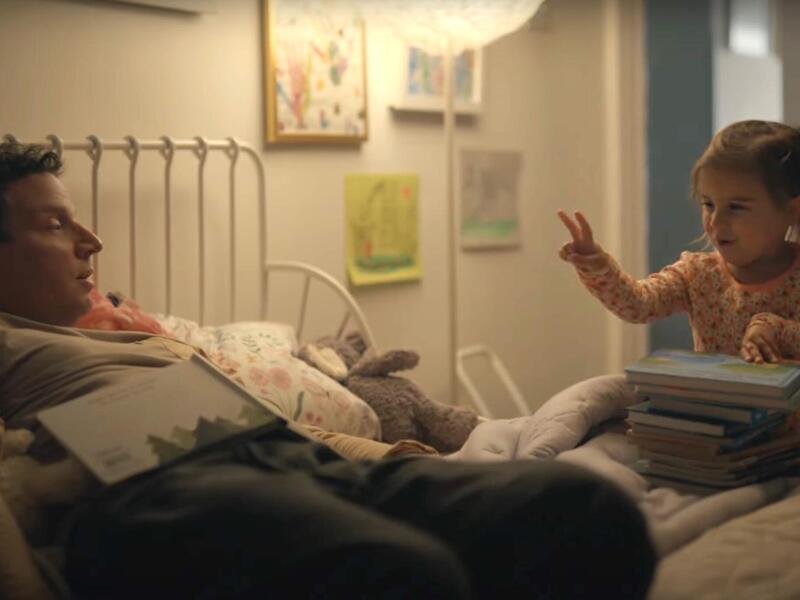 A photo of man laying on a bed. Standing next to the bed is a little girl holding up one hand with two fingers outstretched. 