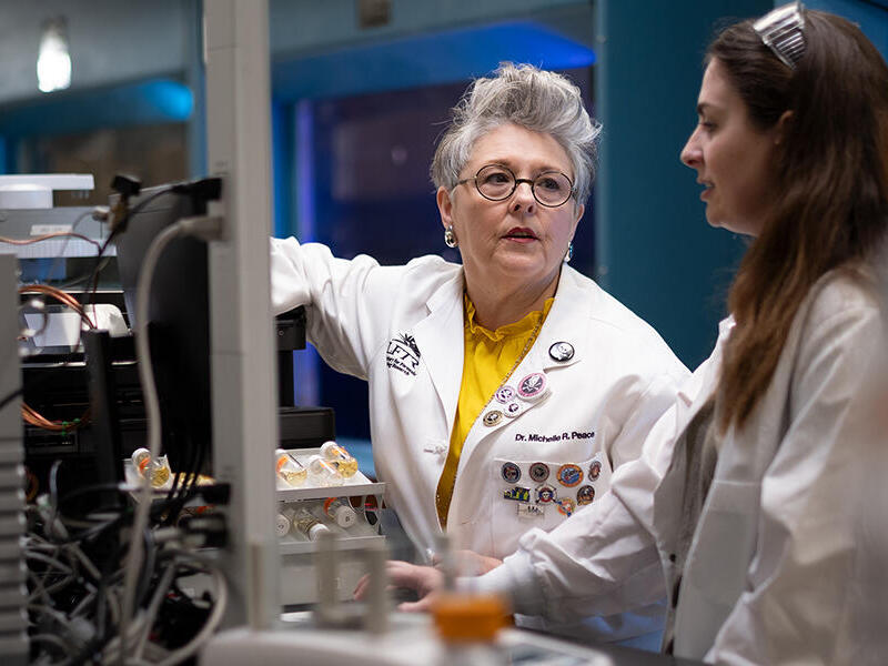 A photo of two women standing in a lab wearing white lab coats. 