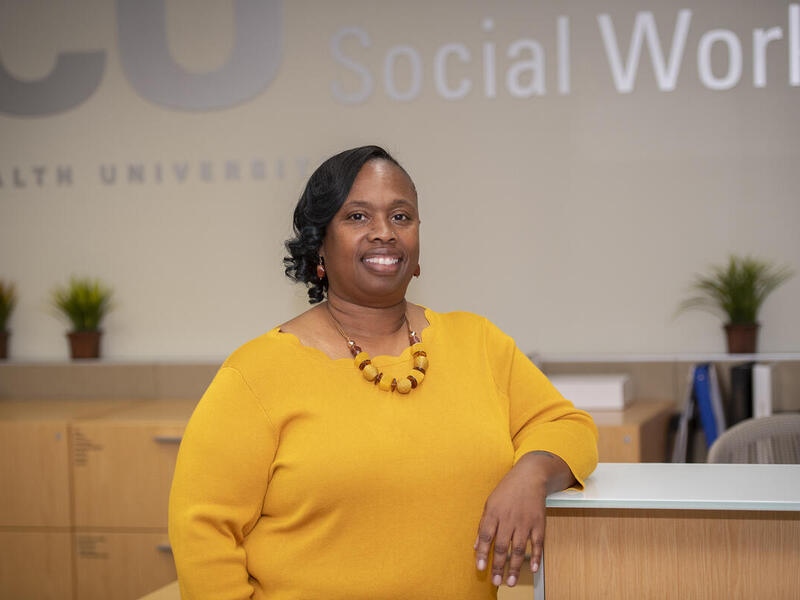 Shenita Williams leaning on a desk in front of a wall that says \"Social Work\" in silver letters. 