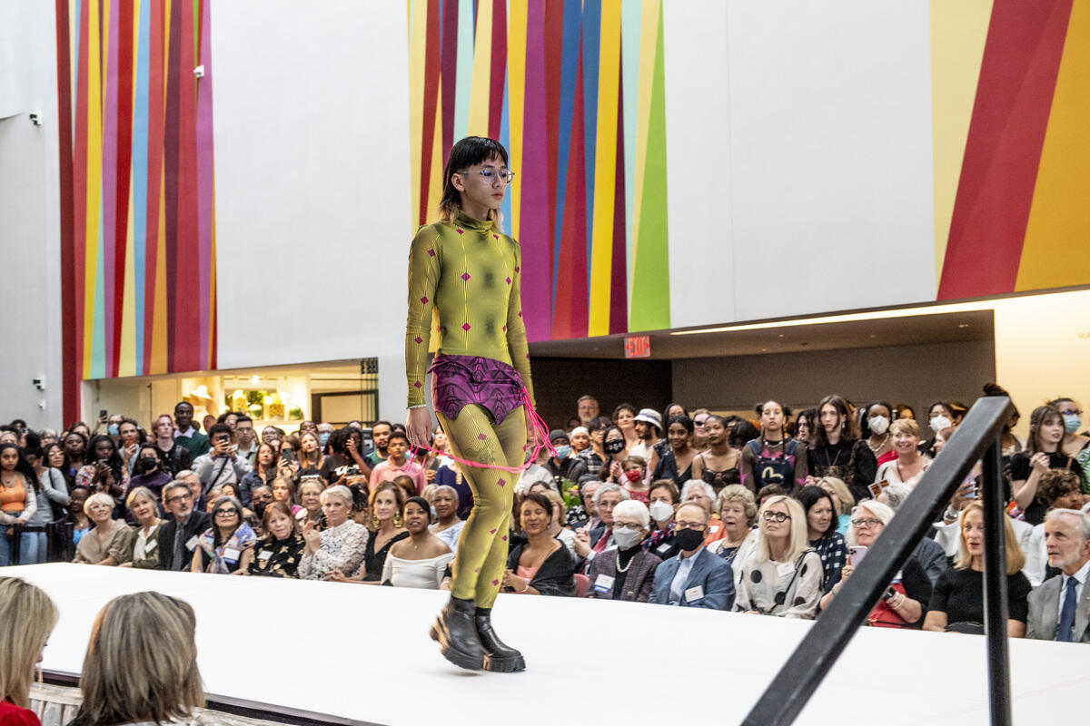 A fashion model walking down a fashion runway surrounded by people at the VMFA. The model has short black hair, glasses and is wearing a patterned greet jumpsuit, purple shorts, and black boots. 