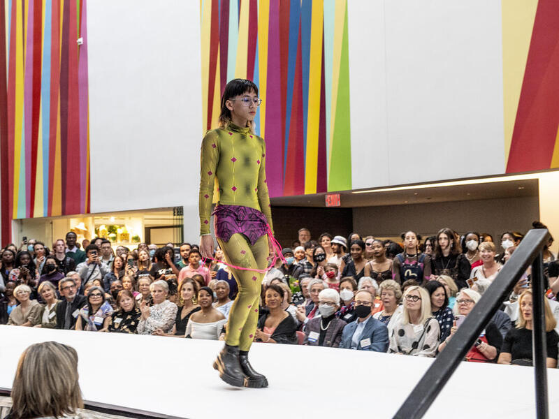 A fashion model walking down a fashion runway surrounded by people at the VMFA. The model has short black hair, glasses and is wearing a patterned greet jumpsuit, purple shorts, and black boots. 