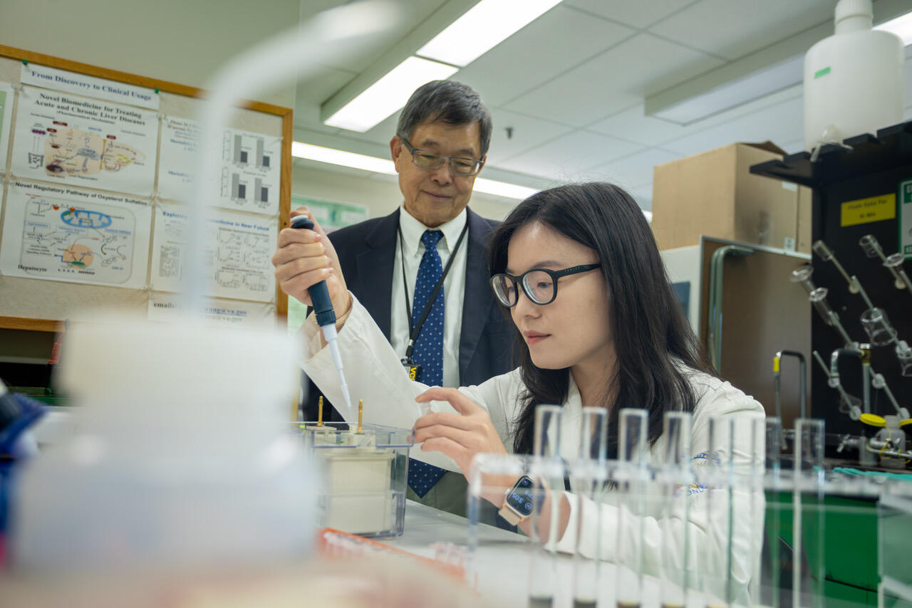 A photo of a man in a suit and tie standing behind a woman sitting at a table. She is using a dropper to put liquid into a beaker. 