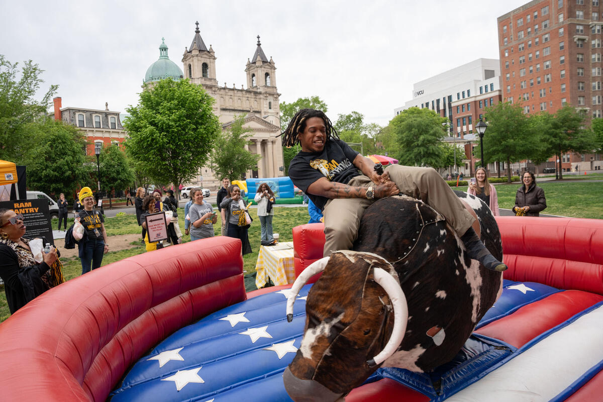 A photo of a man riding a mechanical bull in a park. There is a crowd of people around the bull watching him. 