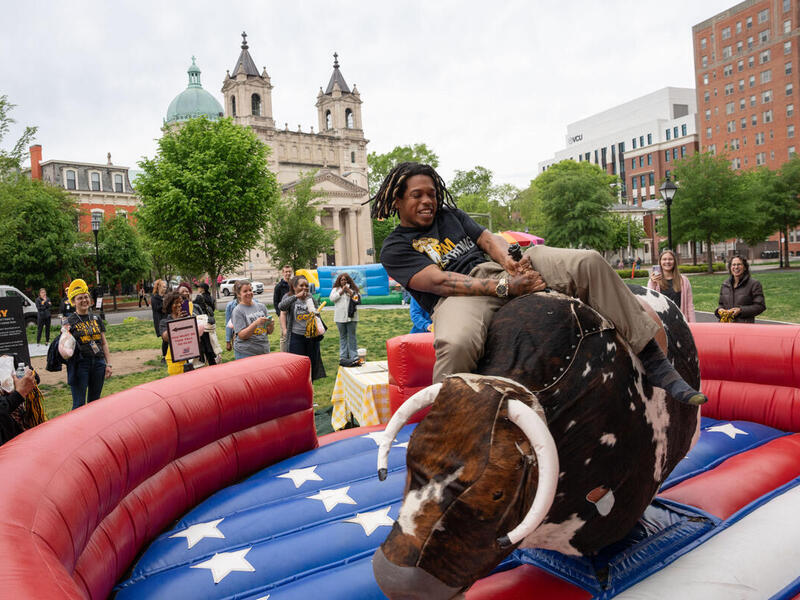A photo of a man riding a mechanical bull in a park. There is a crowd of people around the bull watching him. 