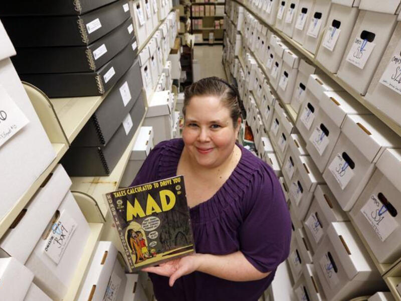 Cindy Jackson holds a rare Mad magazine issue between two rows of shelves of boxes.