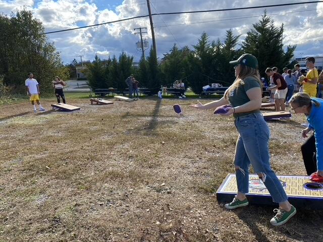 Group of people play cornhole. Woman in the foregroun tosses a bag that is midair.