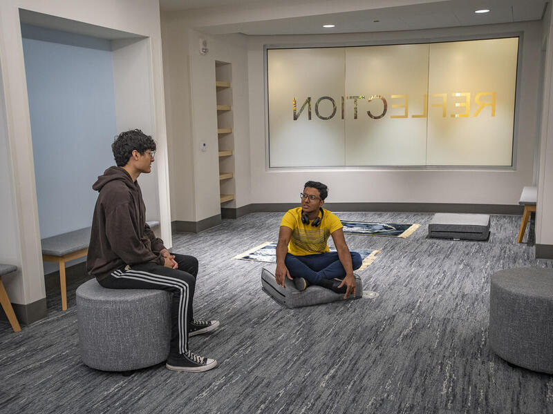 A photo of two men sitting in a room. One is sitting on a stool and the other is sitting on a cushion on the floor. 