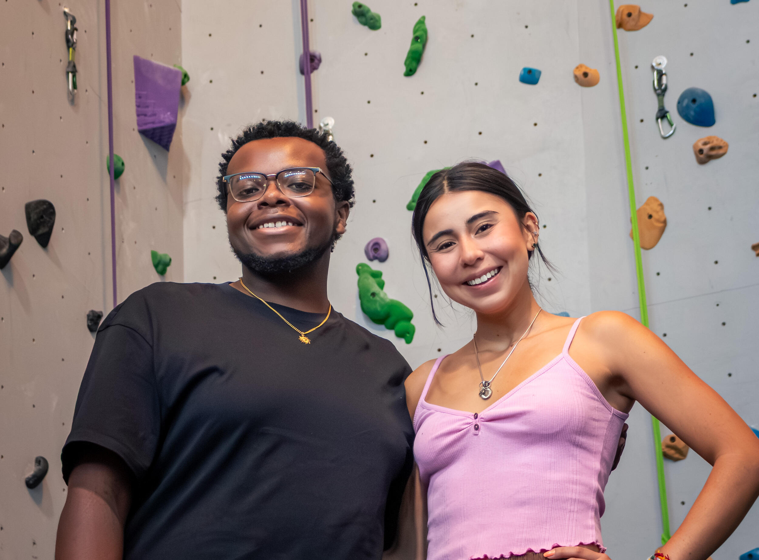Two people stand and smile in front of an indoor rock climbing wall.