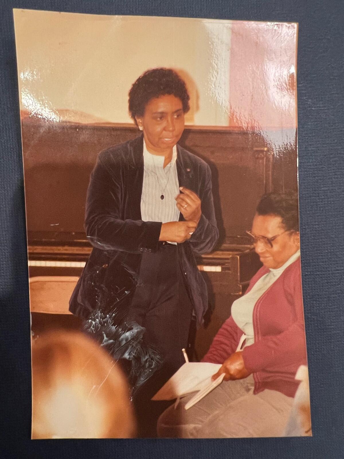 A photo of a woman standng while another woman sits next to her writing down notes. 