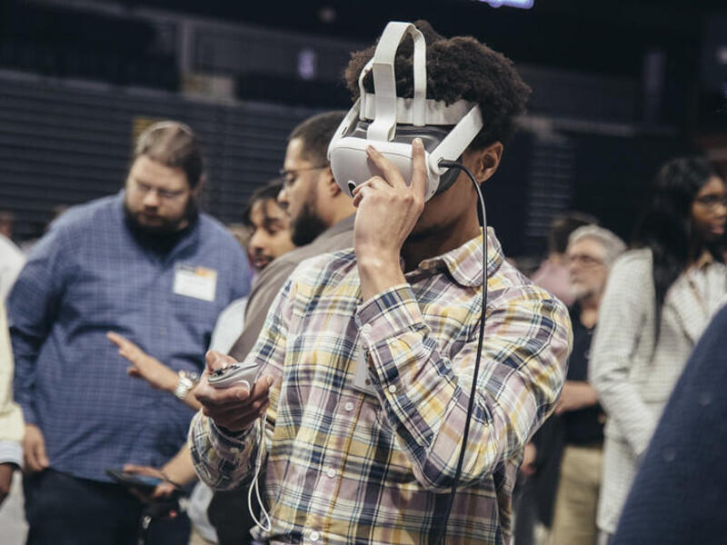 A photo of a man wearing a VR headset and holding a remote. He is surrounded by other people who are walking and looking at other things. 