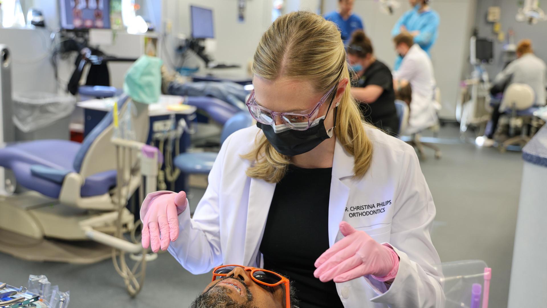 A photo of a woman wearing a lab coat, glasses, and face mask looking down at a man sitting in a dental examination chair. 