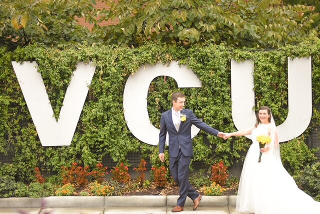A bride and groom hold hands in front of a large VCU sign.