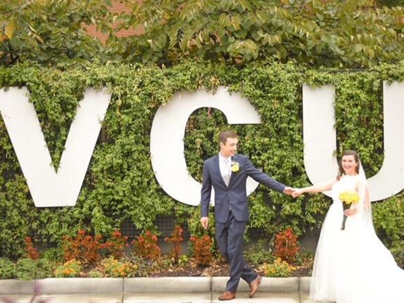 A bride and groom hold hands in front of a large VCU sign.