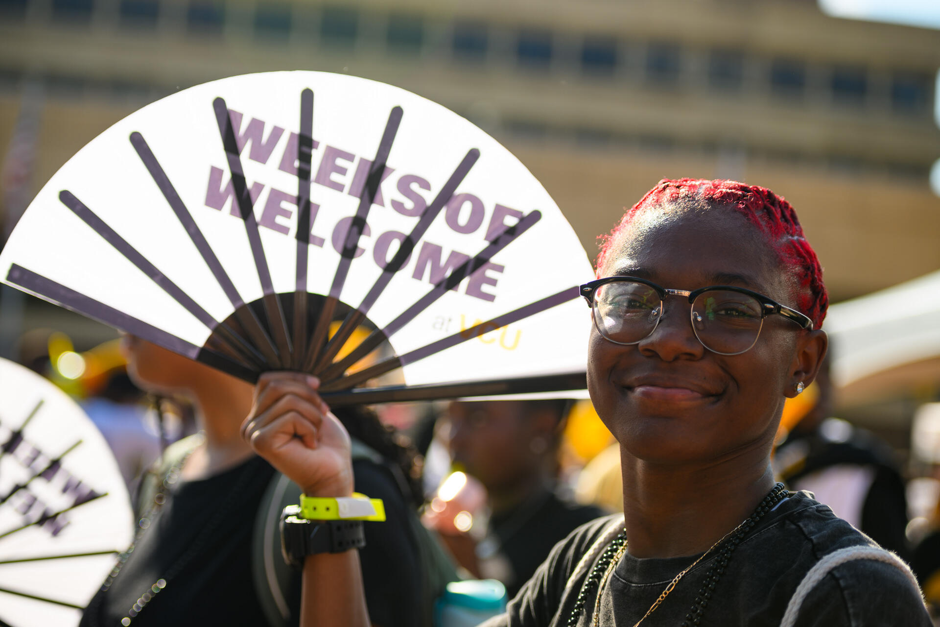 A person in glasses smiles and holds up a fan that says Weeks of Welcome on it.
