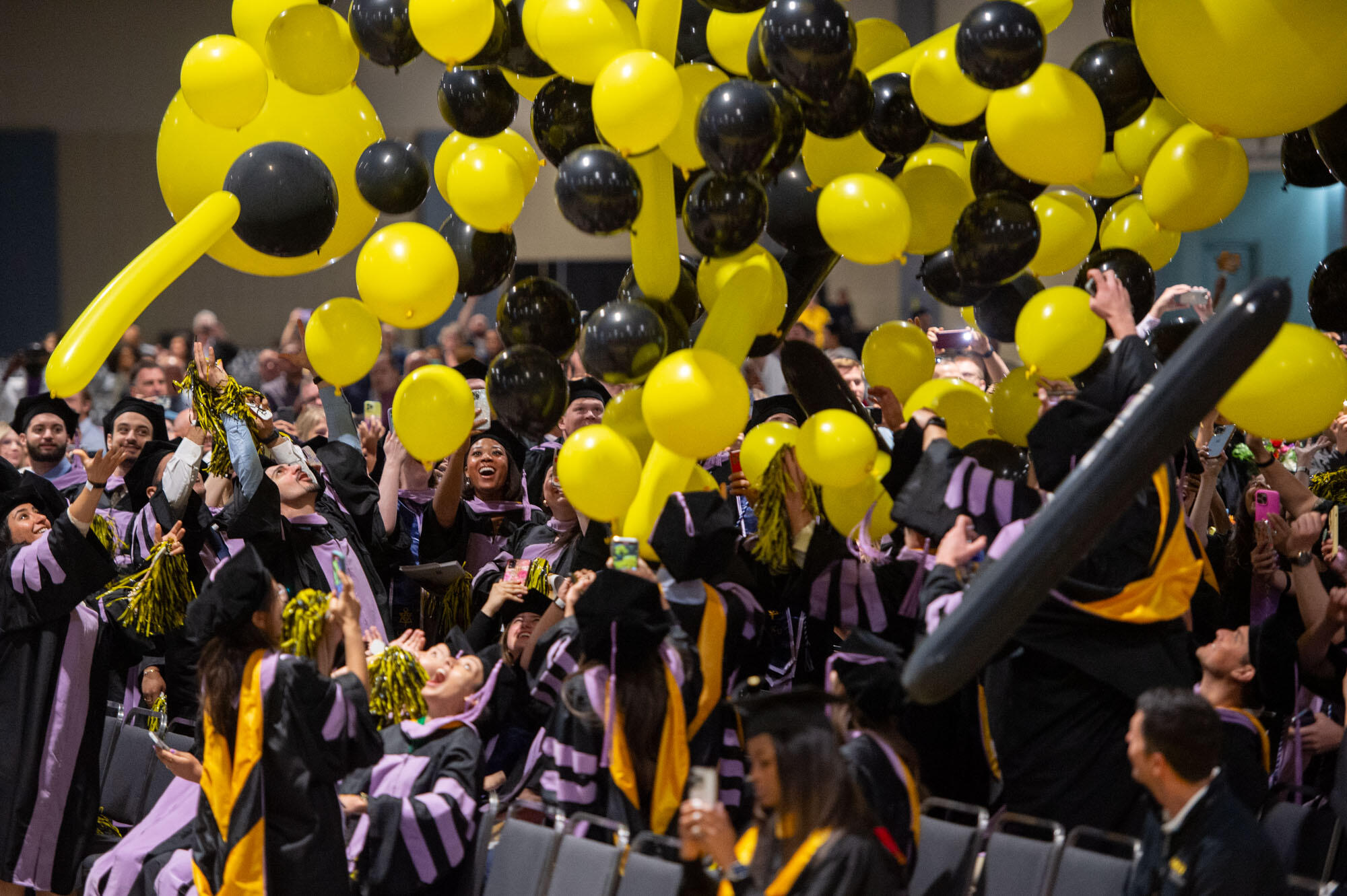 A crowd of people in caps and gowns celebrate with black and yellow balloons above them.