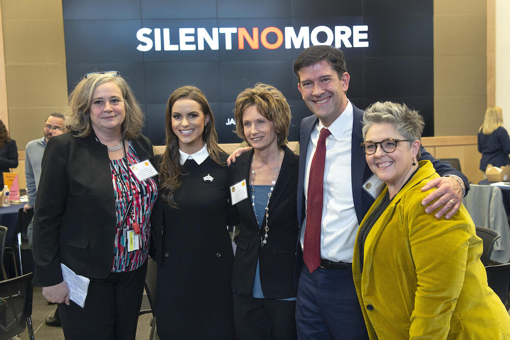 Group stands in front of a sign that reads 'silent no more'