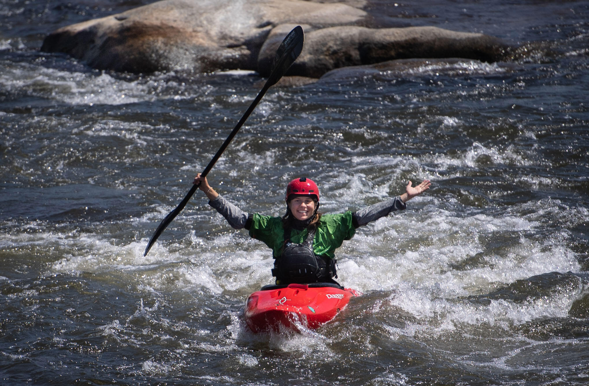 A photo of woman riding in a kayak through rapids. She is holding her arms out to the side in triumph and holding her ore up in her left hand. 