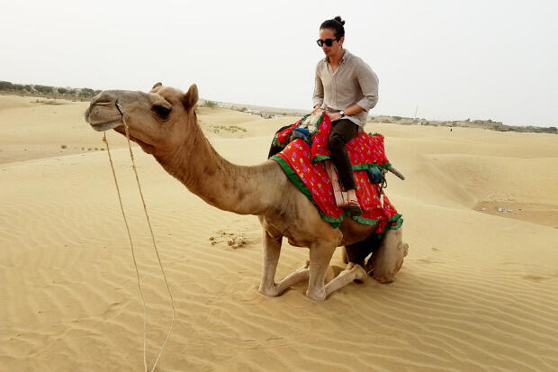 A man sits on a camel with a red saddle in a desert.