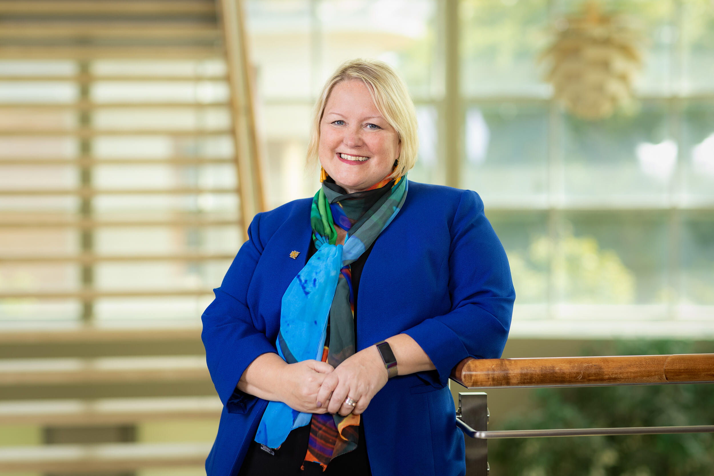 A photo of a woman from the waist up. Her right arm is resting on the railing of an indoor balcony. 