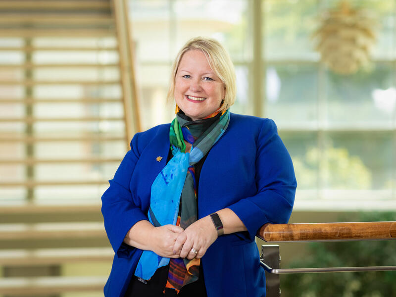A photo of a woman from the waist up. Her right arm is resting on the railing of an indoor balcony. 
