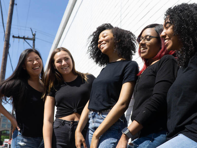 Diverse group of 5 girls, all in black shirts, stand together smiling. 