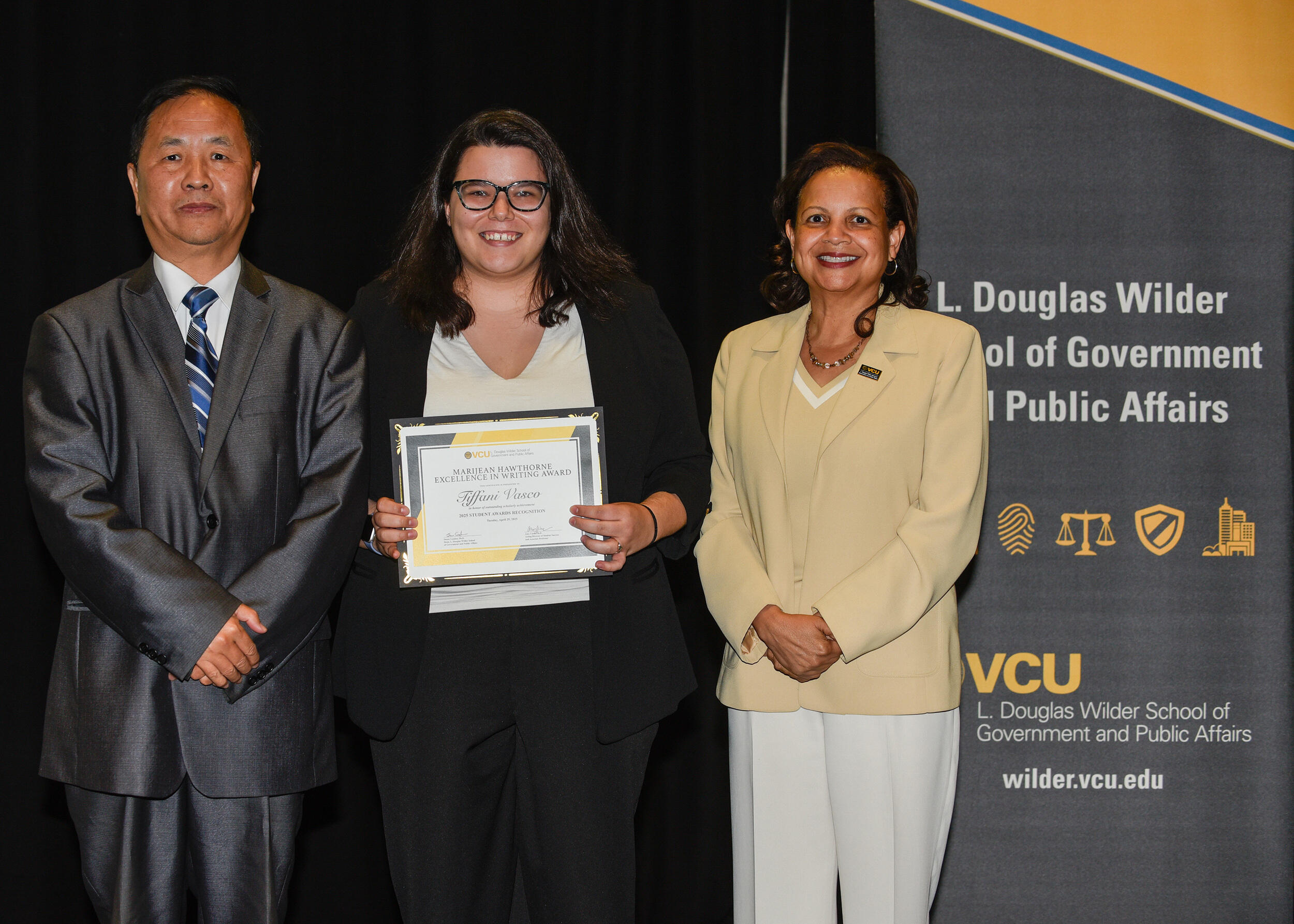A photo of three people standing. The woman in the middle is holding up a certificate. 