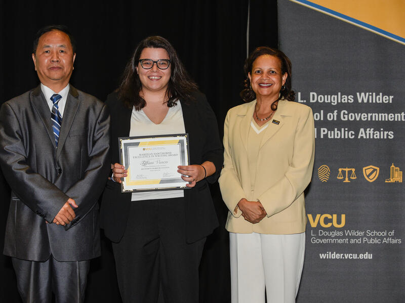A photo of three people standing. The woman in the middle is holding up a certificate. 