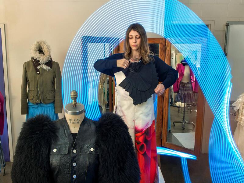 A photo of a woman dressing a maniquin with other maniquins in outfits around her