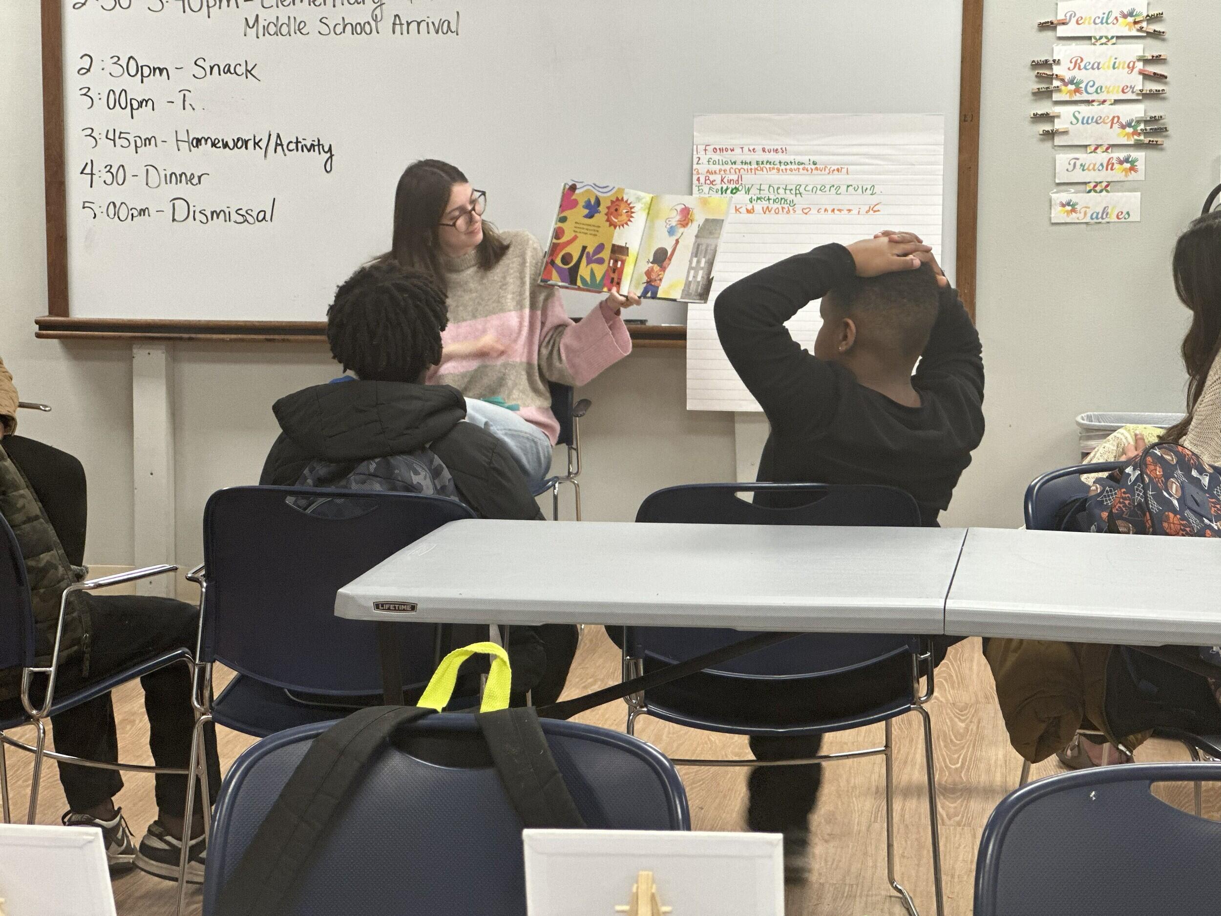 A woman in glasses and a sweater reads from a picture book at the front of a classroom as children watch.