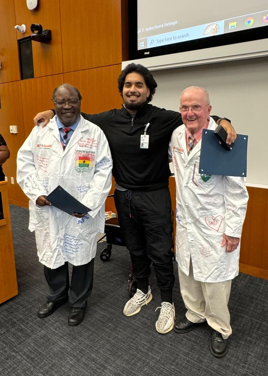 A photo of three men standing. The man in the middle has his lef arm around the shoulder of the man on his left, and his right arm around the shoulders of the man on his right. 