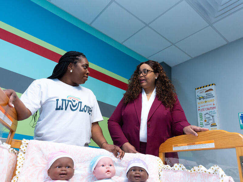Two women facing each other in front of a crib with three baby dolls. 