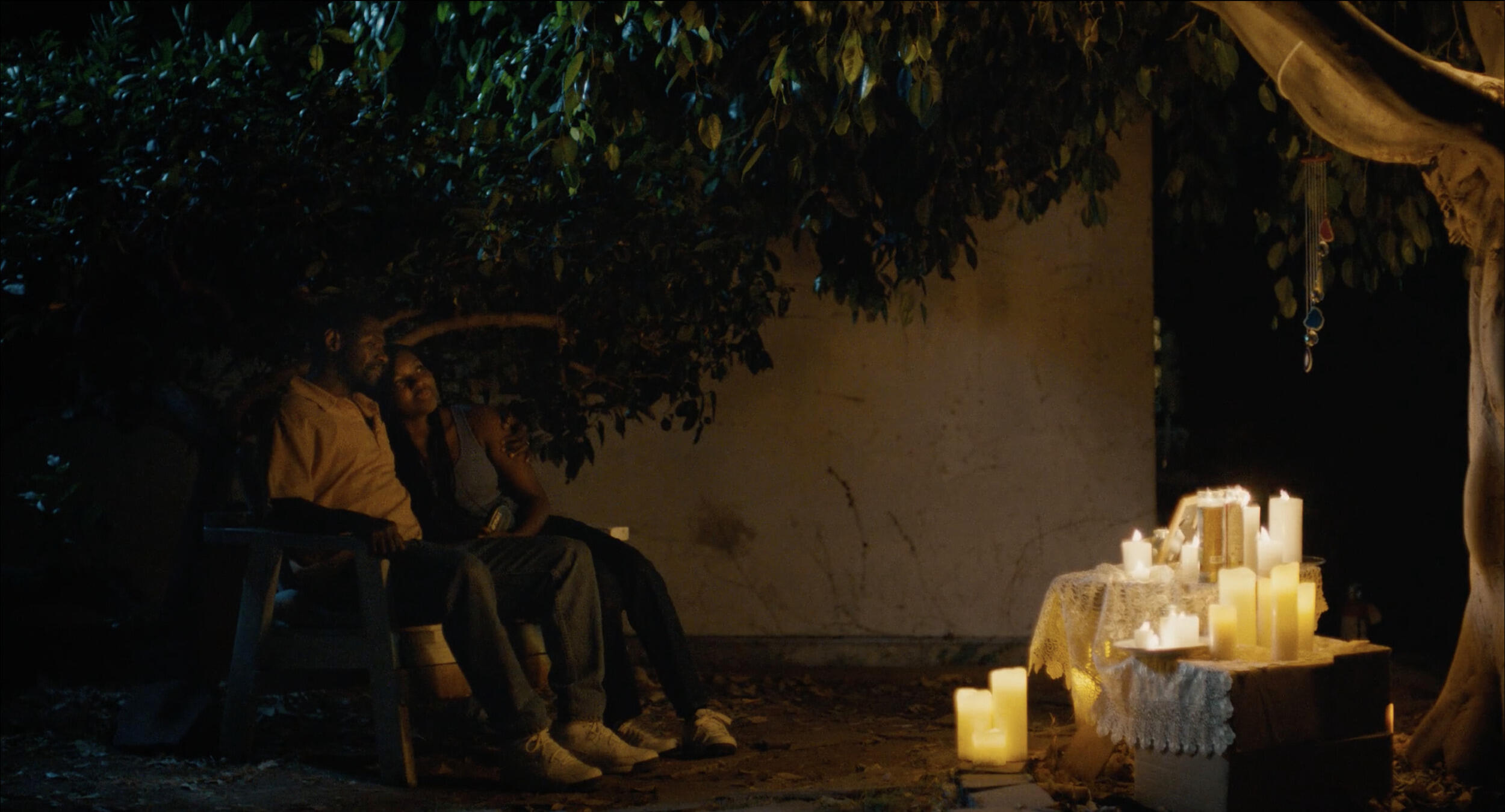 Two people sitting outside during nighttime facing several candles sitting on boxes and the ground. 