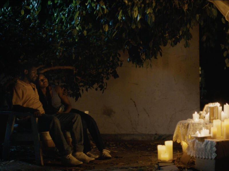 Two people sitting outside during nighttime facing several candles sitting on boxes and the ground. 