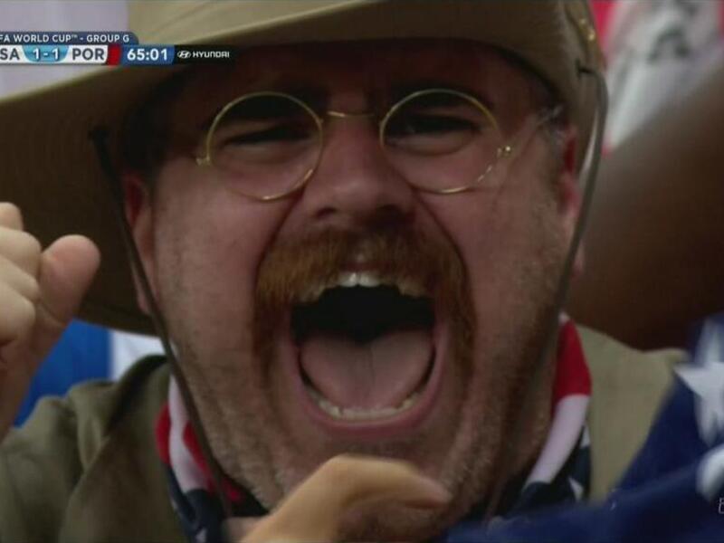 A man in a mustache, glasses and a hat cheers.