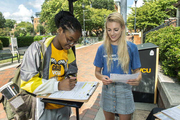 A woman in a yellow T-shirt and gray jacket signs a paper on a clipboard as another woman, in blue shirt, holds another paper and watches.
