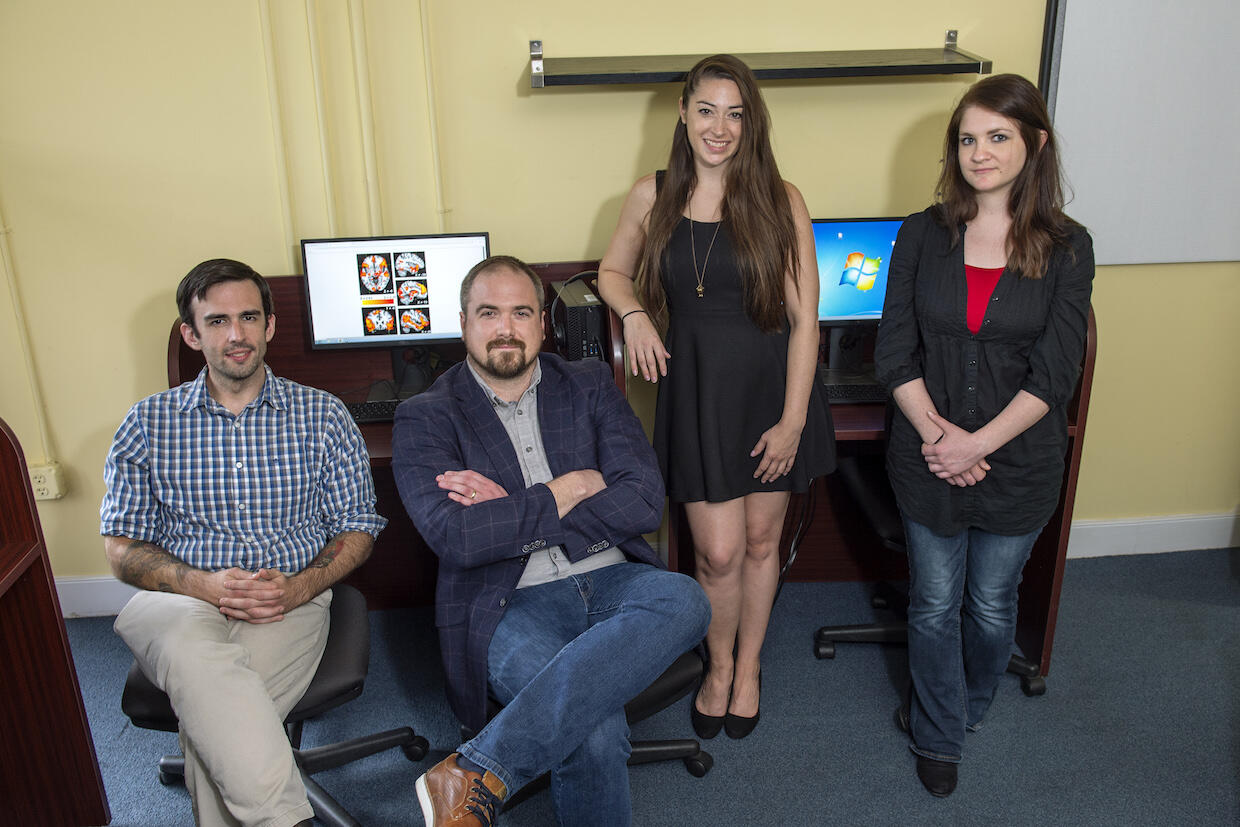 A group of doctoral students posing for a photo. Brain scan imaging is displayed on the computer screen behind the two left figures.
