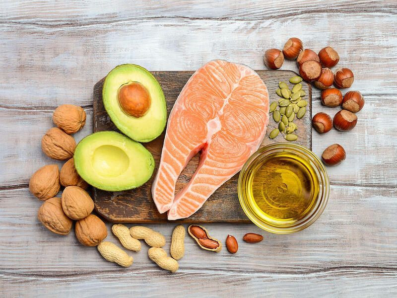 A cutting board with a cut of salmon, a split avocado, seeds and a cup of olive oil on it with hazelnuts, peanuts and walnuts around its sides.