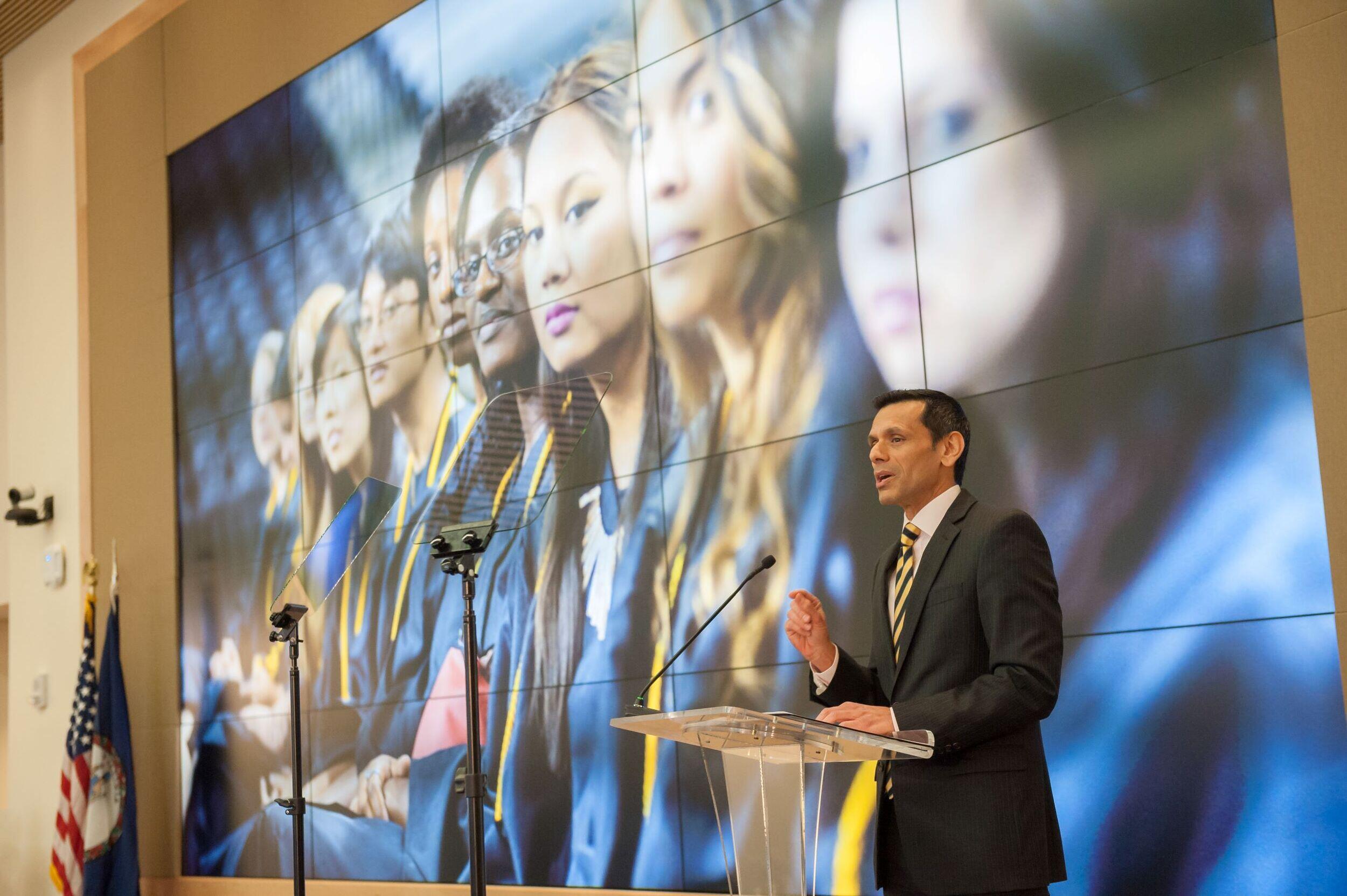 Photo of Michael Rao speaking at a podium with a large image behind him of a group of people looking toward the camera.
