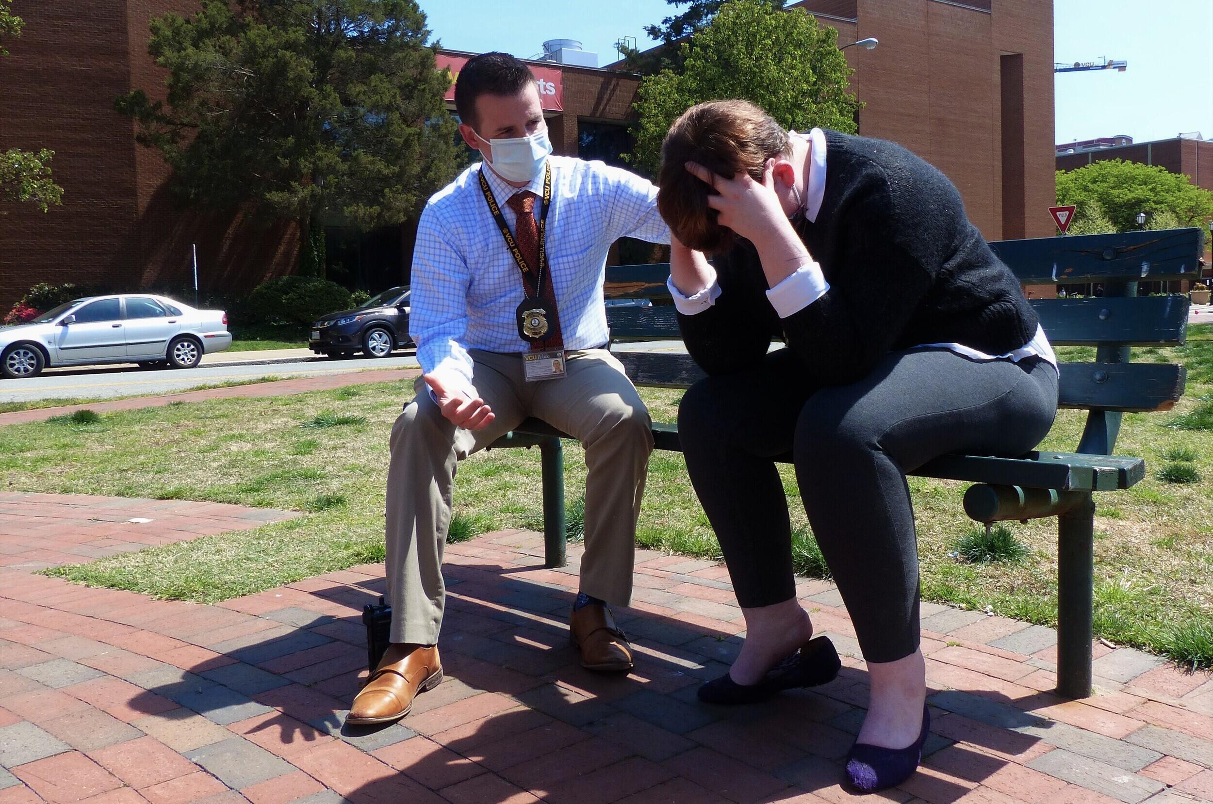 Two people seated on a bench — one person speaks to another with their head in their hands.