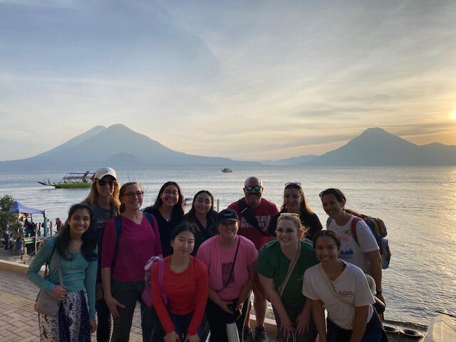 A group photo of 12 people standing in front of a lake near some mountains. 