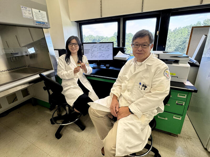 A woman and man in lab coats sit on chairs. The woman holds up a bottle.