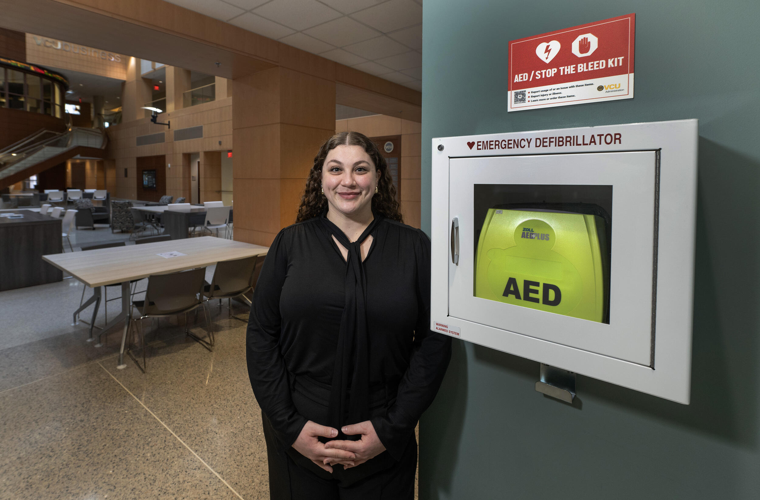 A photo of a woman standing next to an emergency defibrillator box that is hanging on a wall.
