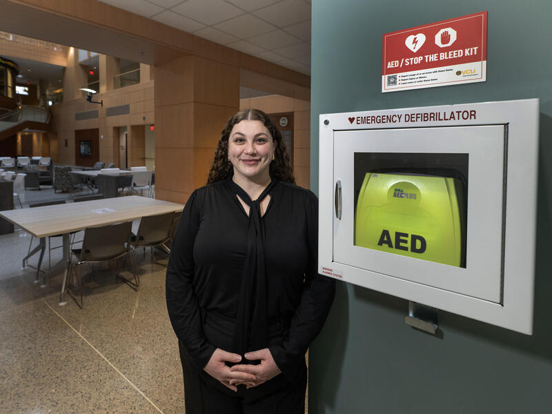 A photo of a woman standing next to an emergency defibrillator box that is hanging on a wall.