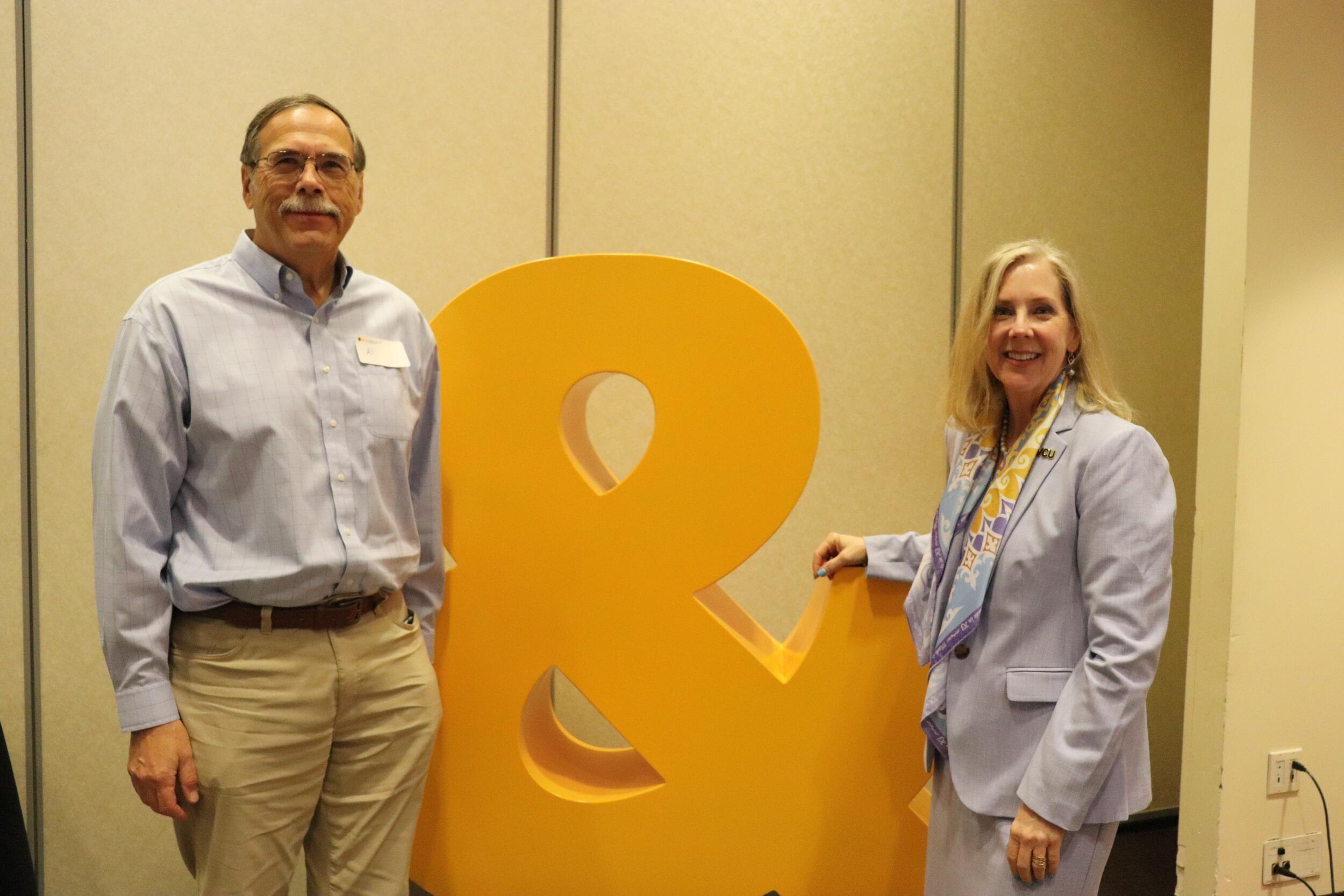 A man and woman standing on either side of a large yellow ampersand statue. 