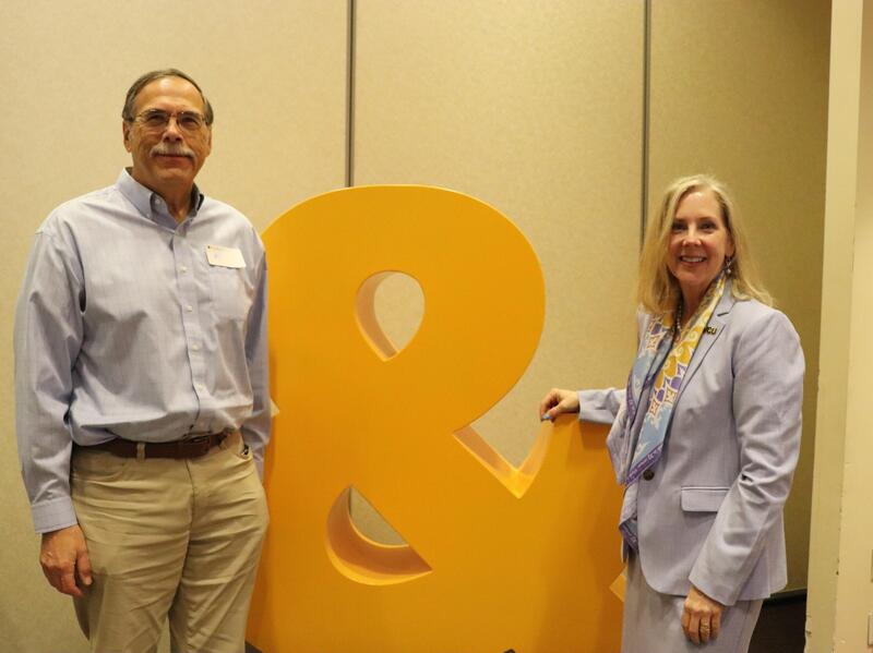 A man and woman standing on either side of a large yellow ampersand statue. 