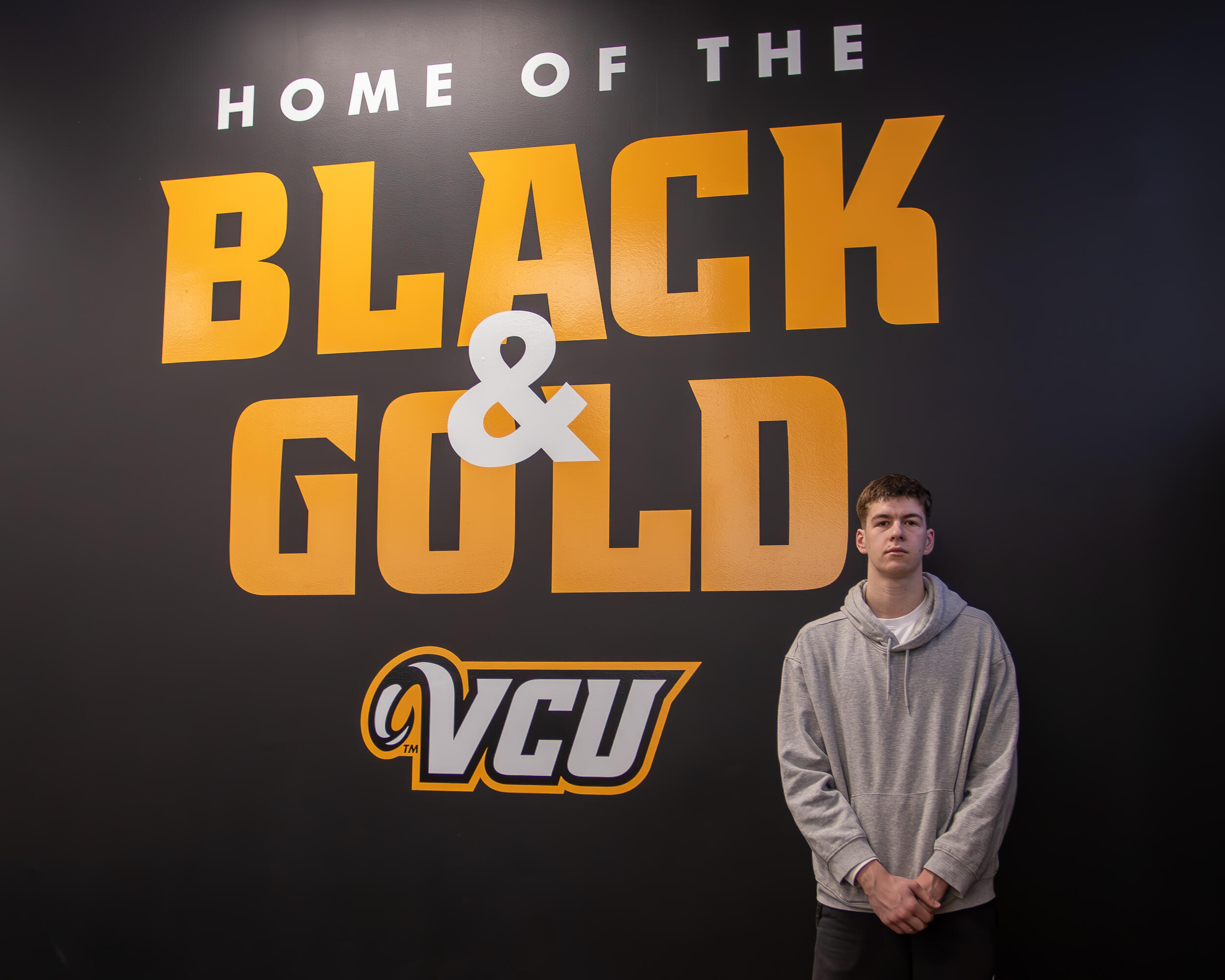 A photo of a man standing in front of a back whall with white and yellow letters that spell out \"HOME OF THE BLACK & GOLD VCU\" 