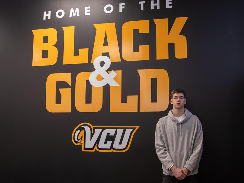 A photo of a man standing in front of a back whall with white and yellow letters that spell out \"HOME OF THE BLACK & GOLD VCU\" 