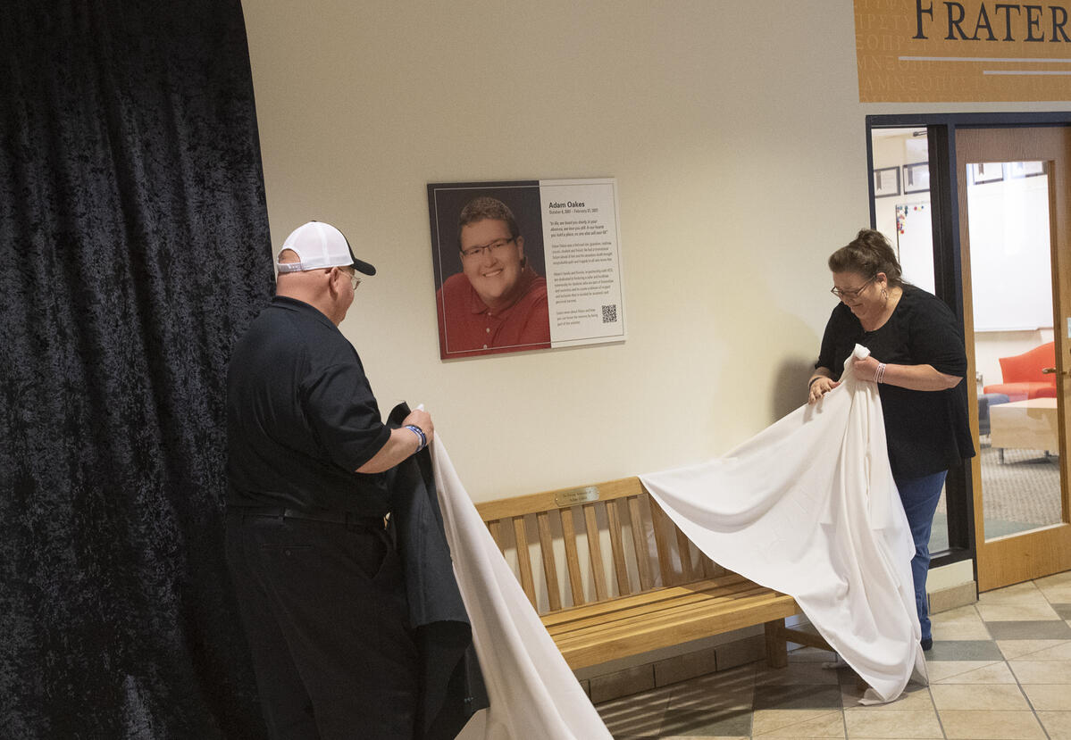 A man and a woman standing on either side of an image of Adam Oaks. The woman is holding a white cloth and crying. 