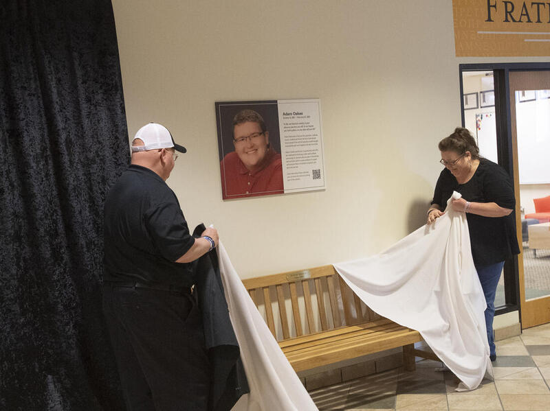 A man and a woman standing on either side of an image of Adam Oaks. The woman is holding a white cloth and crying. 